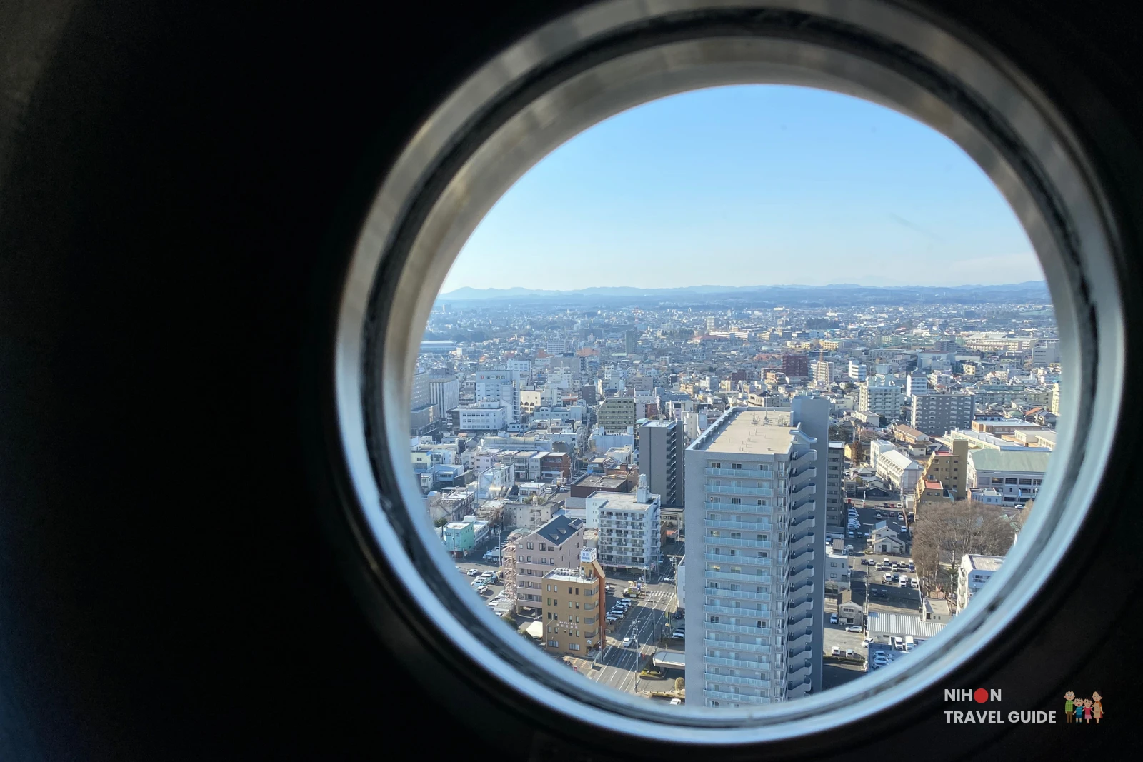 View through a circular porthole window on the Art Tower Mito observation deck, looking west over Mito City's urban skyline with a tall residential tower in the foreground and the faint twin-peaked silhouette of Mount Tsukuba (877m) visible on the distant horizon, Mito City, Ibaraki, Japan