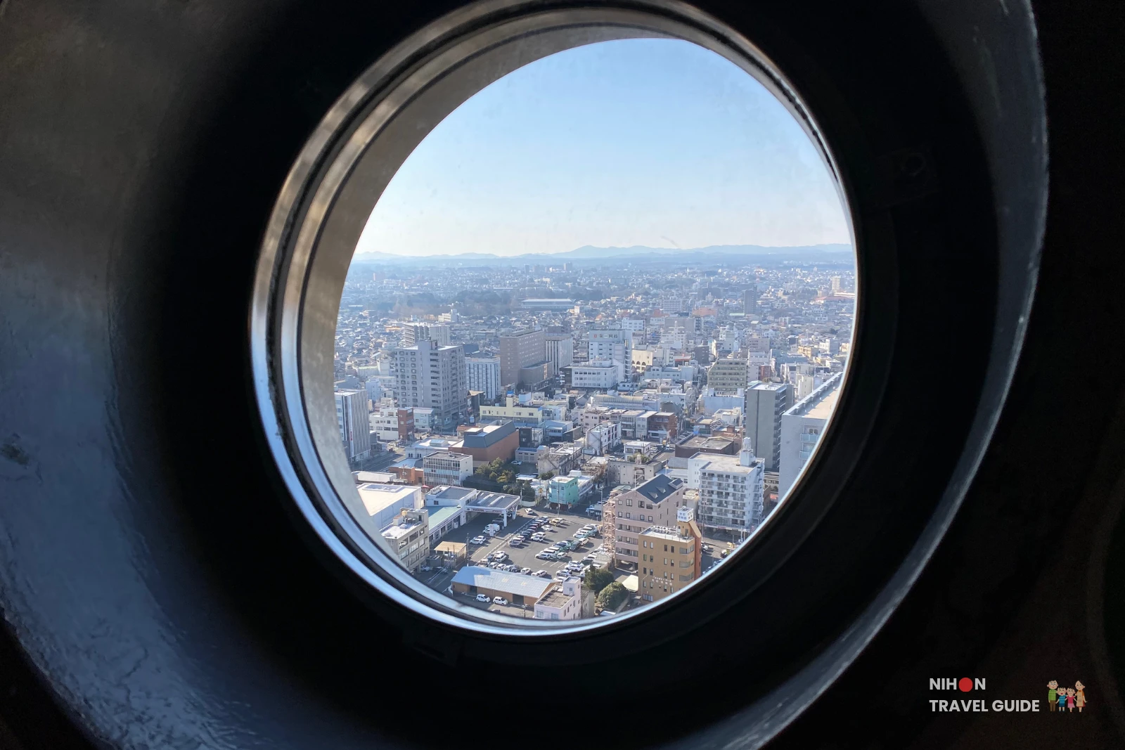 View through a circular porthole on the Art Tower Mito observation deck showing the spread of Mito City below and the distinctive twin-peaked silhouette of Mount Tsukuba (877m, "the Fuji of the East") clearly visible as a low mountain ridge on the western horizon, Ibaraki, Japan