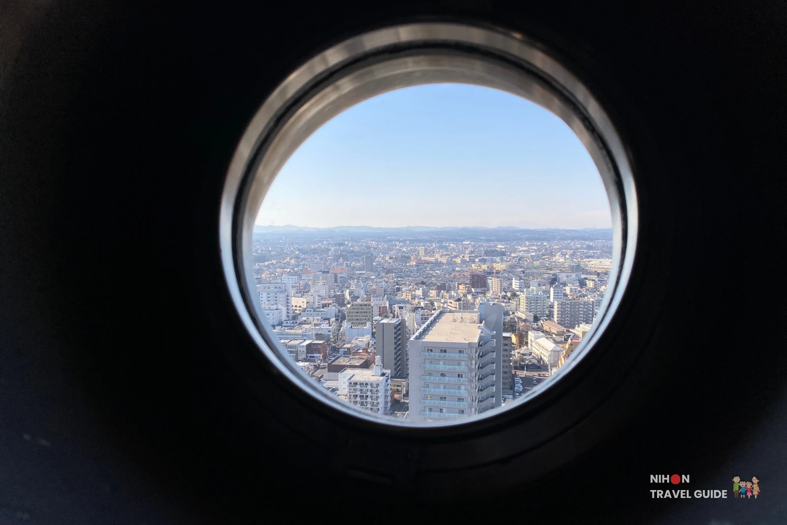 Panoramic view of western Mito City through a circular porthole on the Art Tower Mito observation deck at 86.4 metres, showing a dense urban landscape of low-rise buildings stretching to the horizon with the twin peaks of Mount Tsukuba faintly visible in the haze on the far left, Ibaraki, Japan