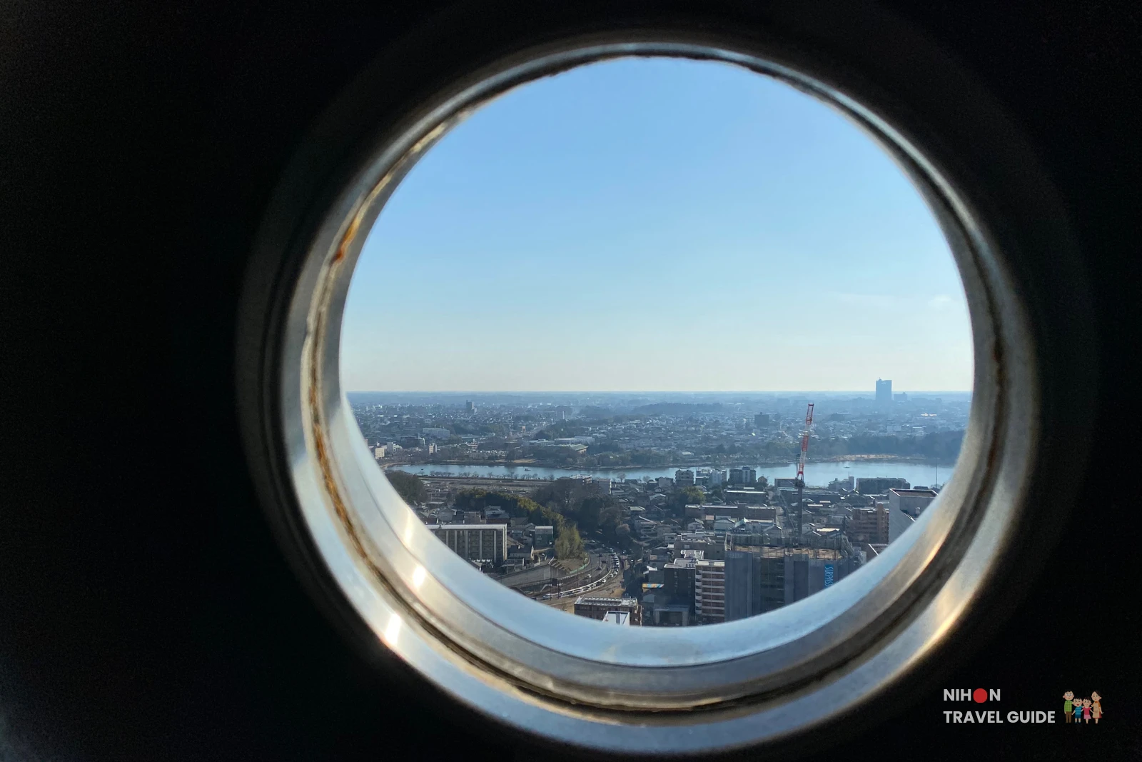 View of Mito City and the Naka River from a circular porthole window on the Art Tower Mito observation deck at 86.4 metres, showing the city spreading to the horizon under a clear blue sky, with a construction crane and the Ibaraki Prefectural Government building visible in the distance, Ibaraki, Japan