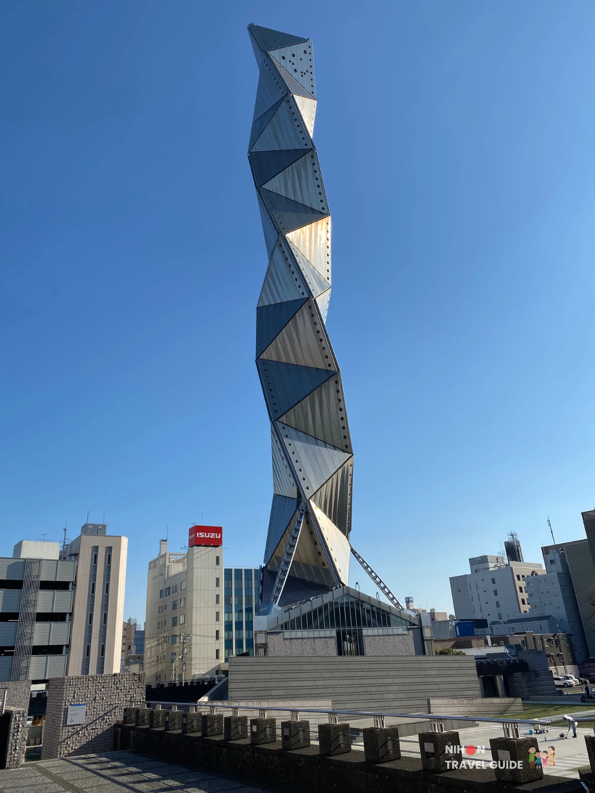 Art Tower Mito's 100-metre titanium spiral tower viewed from the second floor of the complex, showing the full triple-helix structure rising above the stepped stone plaza and surrounding Mito City buildings including an Isuzu dealership sign, under a clear blue sky, Mito City, Ibaraki, Japan