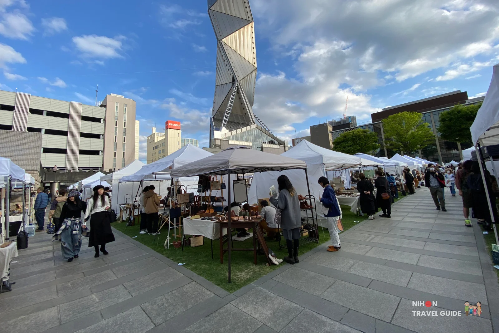Iconic twisted Art Tower Mito rising above white market tents and shoppers at the Aozora Arts & Craft Stall Market.