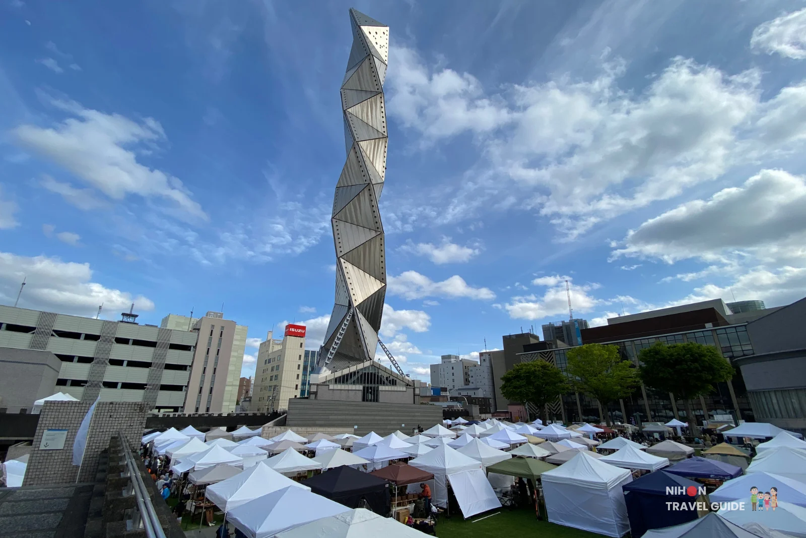 Striking low-angle view of Art Tower Mito’s twisted metallic structure rising into a blue sky, with the bustling Aozora Arts & Craft Stall Market tents filling the foreground.