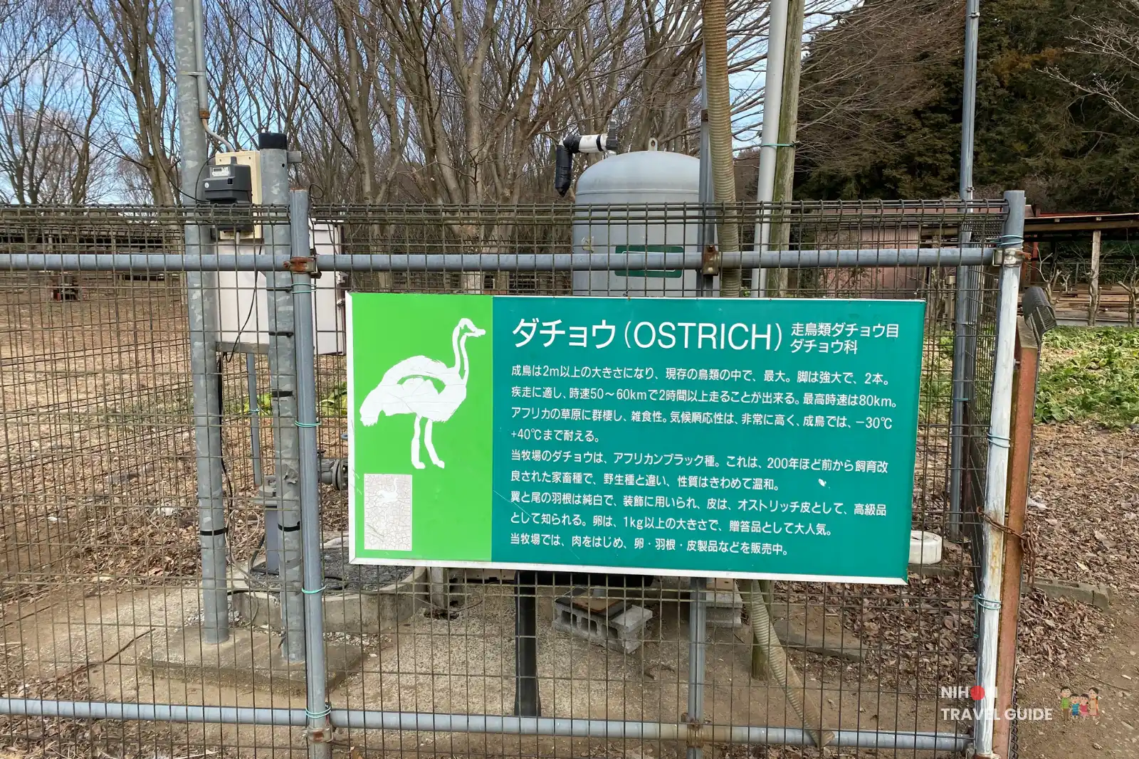 Green information board about ostriches on a fence at Ishioka Ostrich Farm with trees and enclosures in the background