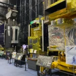 Row of full-scale JAXA Earth observation and communications satellites on display inside the Space Dome at Tsukuba Space Center with visitors walking along the exhibit.
