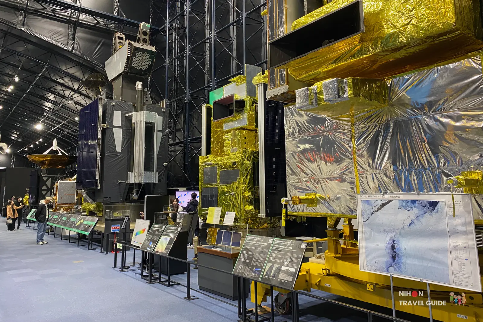 Row of full-scale JAXA Earth observation and communications satellites on display inside the Space Dome at Tsukuba Space Center with visitors walking along the exhibit.