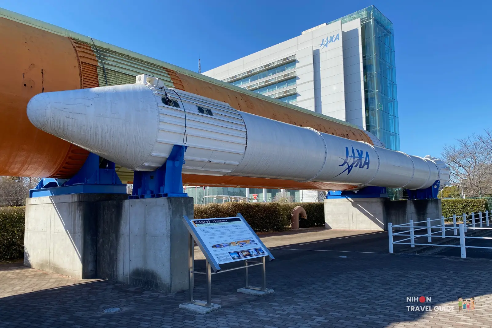Side view of the full-scale H-II rocket display with an information signboard in front and the JAXA Tsukuba Space Center building in the background.