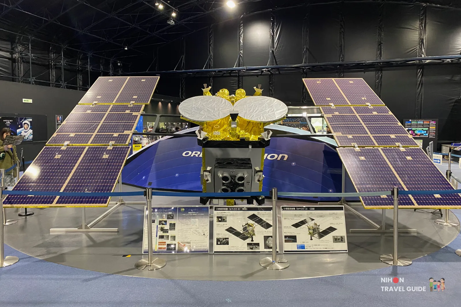 jaxa-tsukuba-hayabusa2-asteroid-sample-return-spacecraft Full-scale Hayabusa2 asteroid sample-return spacecraft display at JAXA Tsukuba Space Center with large solar panel wings extended.