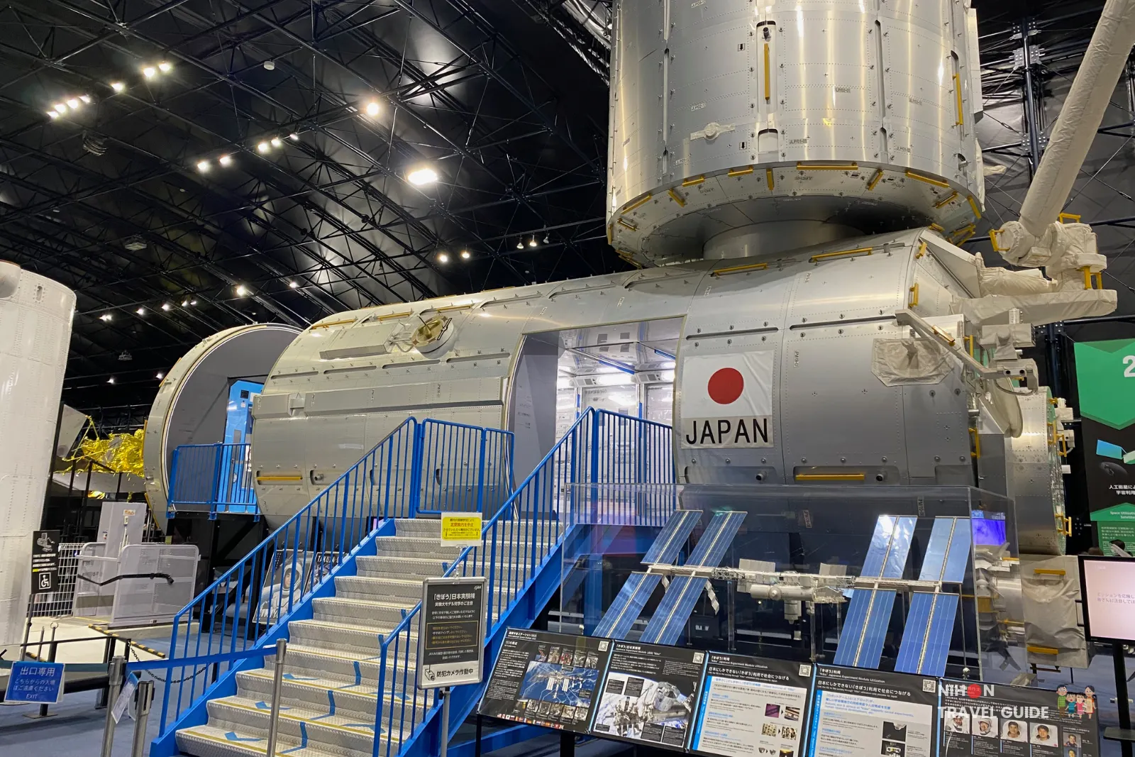Full-scale mock-up of the JAXA Kibō ISS module at Tsukuba Space Center with blue access stairs and an information display below.