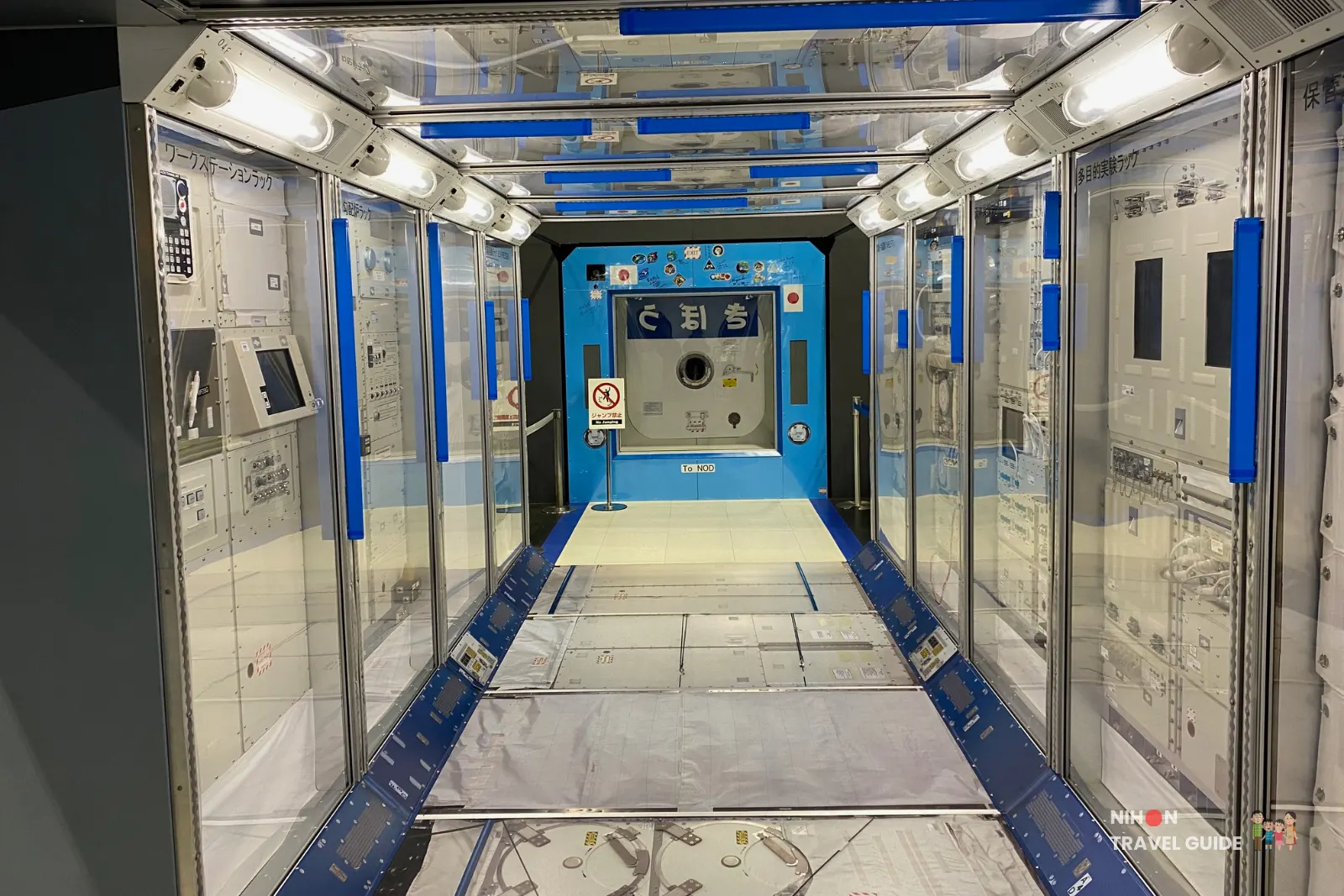 Walk-through interior corridor of the JAXA Kibō experiment module replica with equipment racks lining both sides and a hatch at the far end.