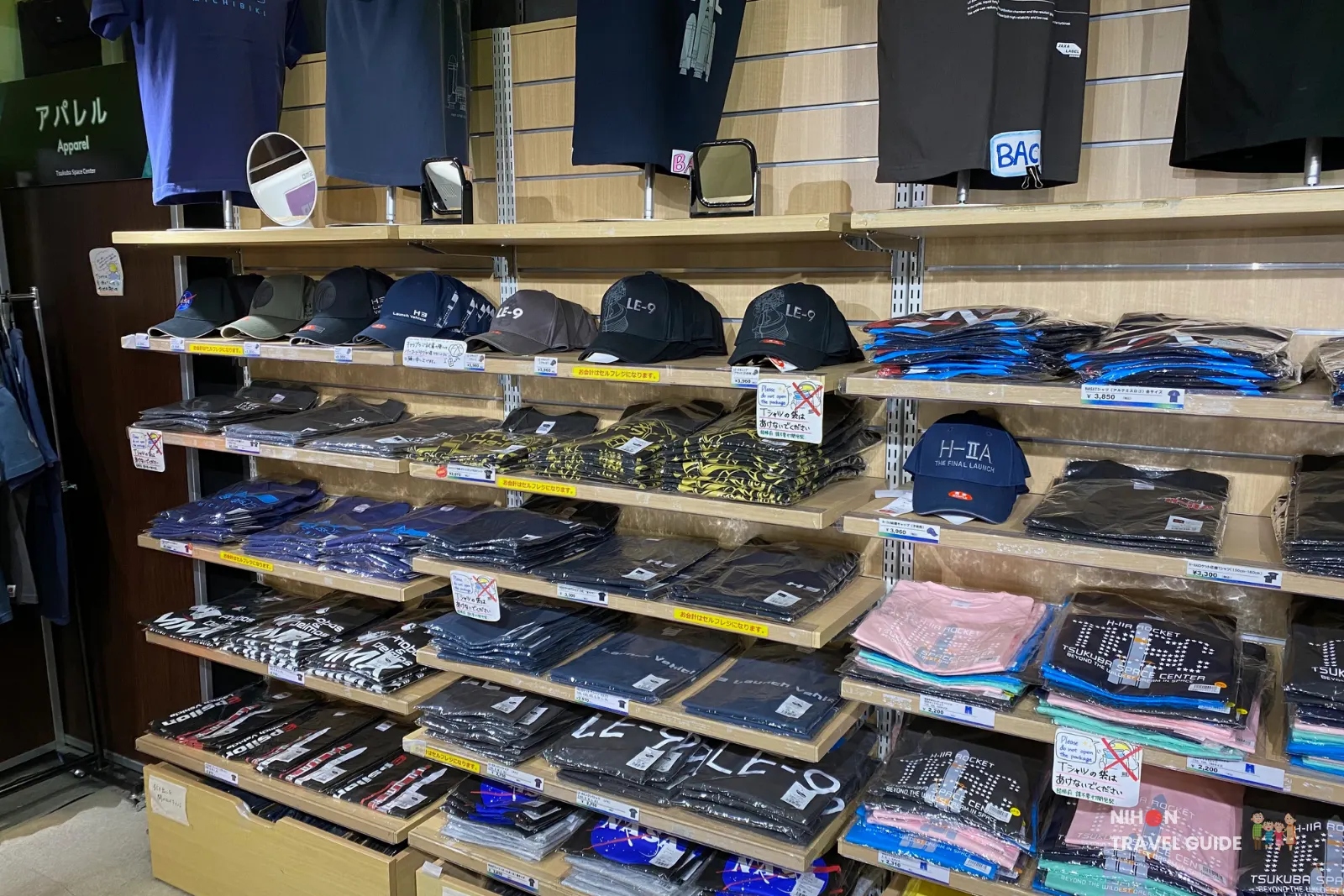 Shelves of JAXA-branded apparel at Planet Cube gift shop with caps and neatly folded T-shirts featuring rocket and mission designs.