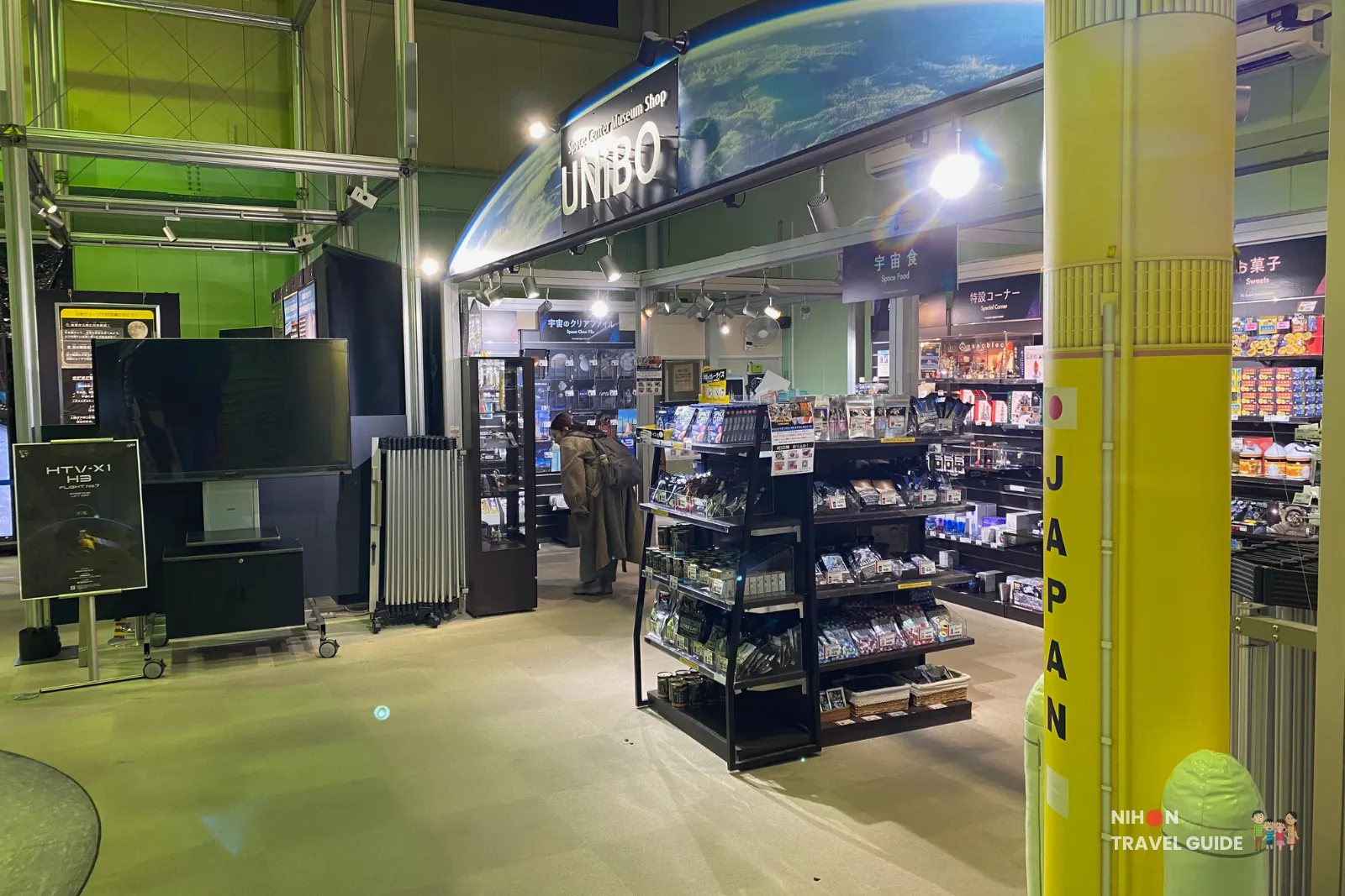 Interior of Planet Cube gift shop at JAXA Tsukuba Space Center with shelves of space-themed souvenirs and a tall yellow rocket pillar near the entrance.