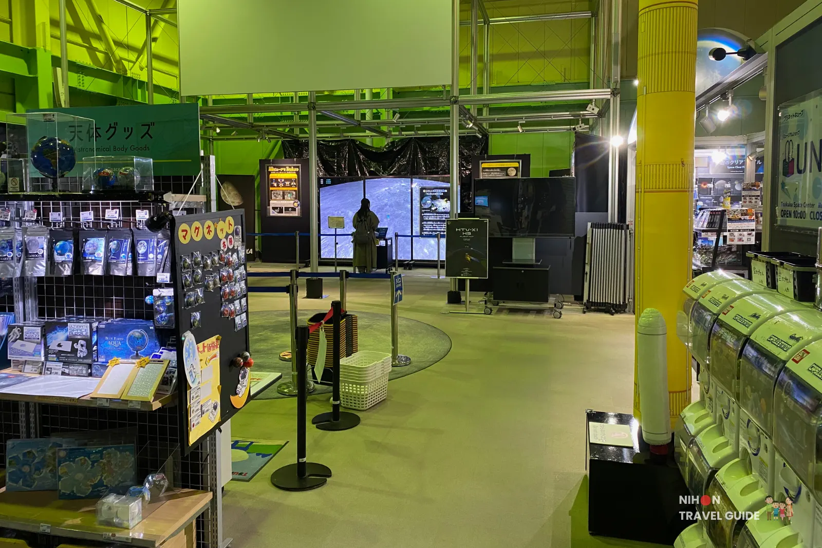 Wide view of the Planet Cube shop area at JAXA Tsukuba Space Center with capsule toy machines, exhibit displays and a central circular floor graphic.