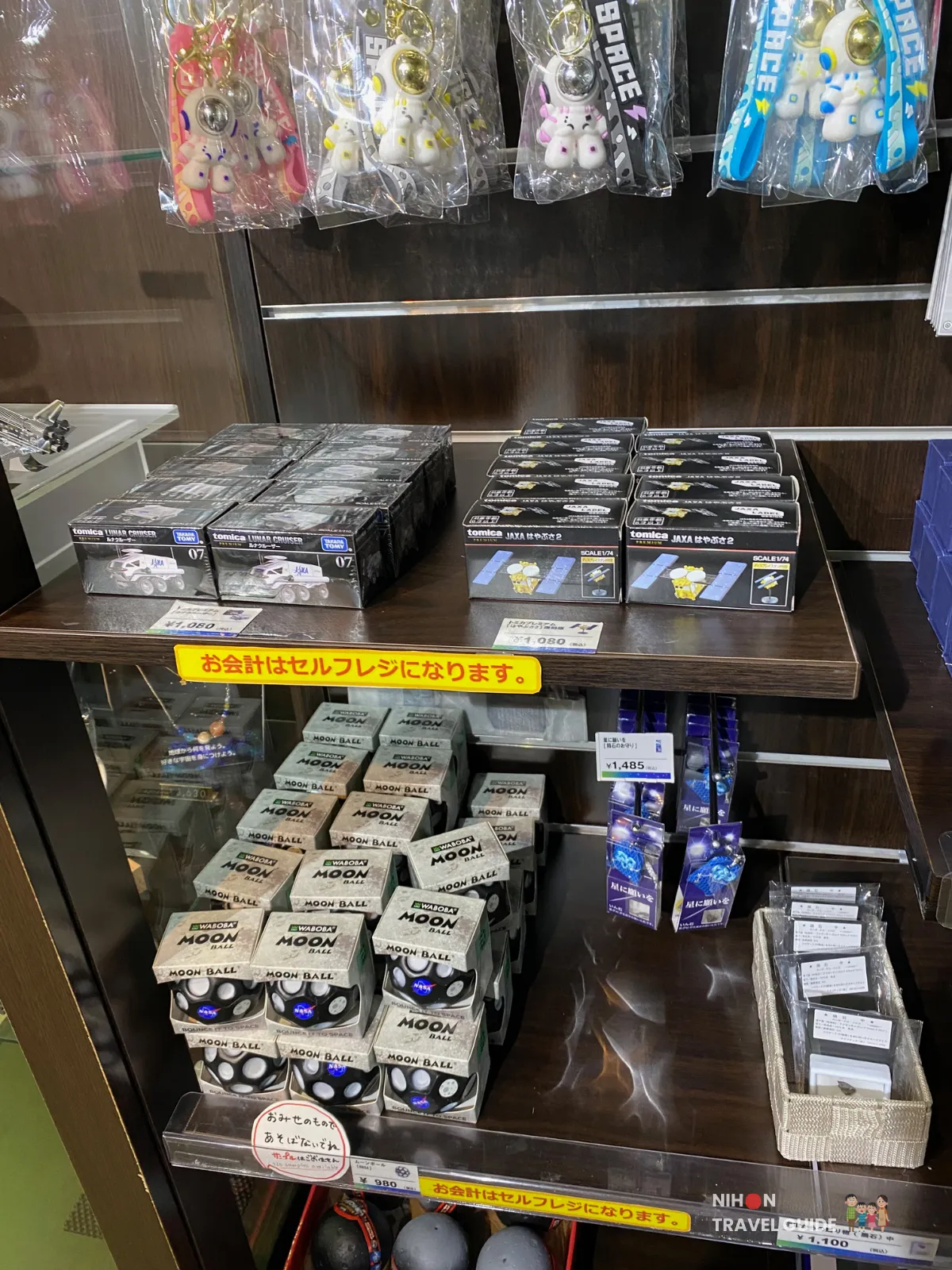 jaxa-tsukuba-planet-cube-souvenir-shelf Merchandise shelf in the Planet Cube gift shop at JAXA Tsukuba Space Center displaying Tomica JAXA Lunar Cruiser toys, moon balls and space-themed keychains.
