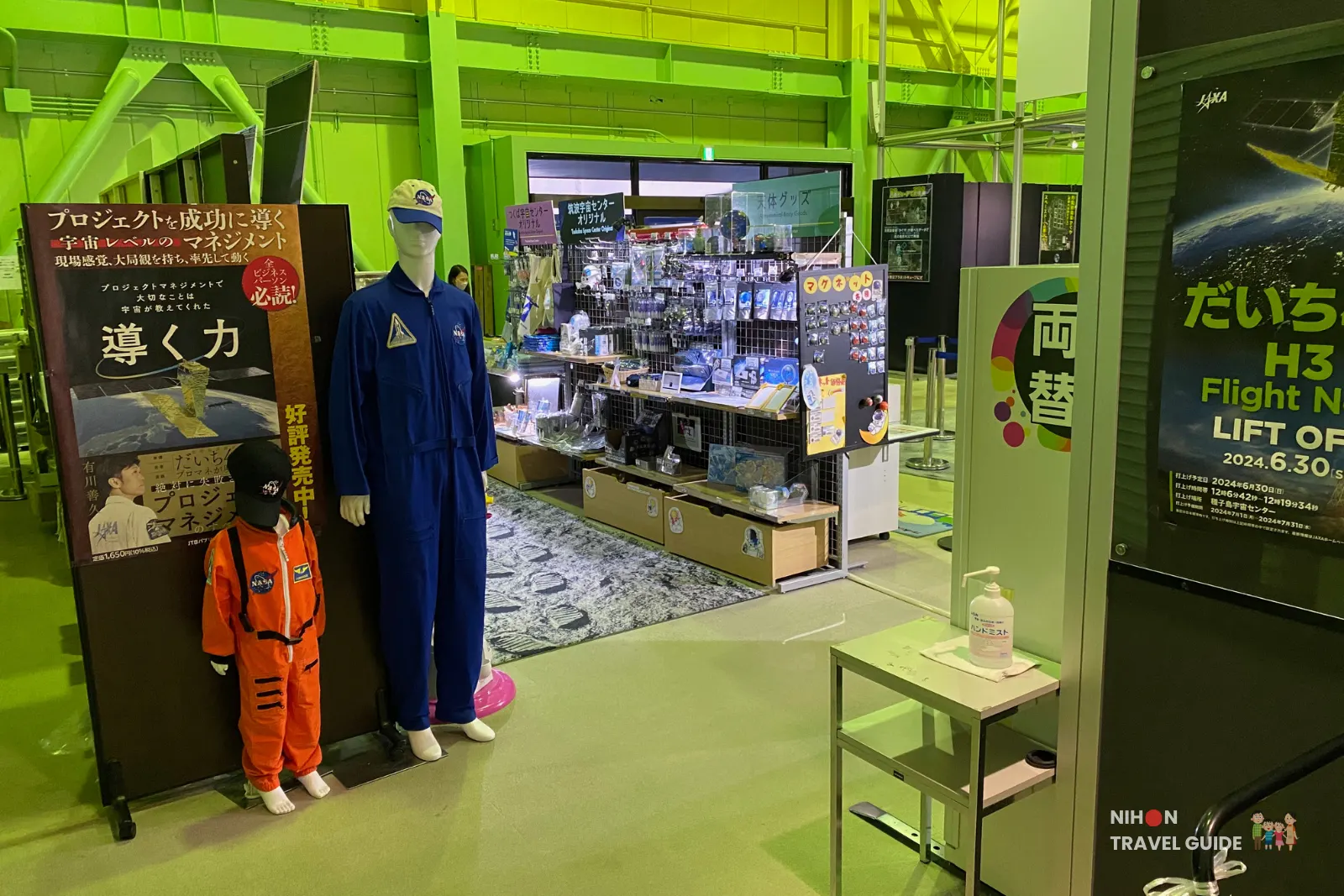 Adult and child spacesuit mannequins standing beside shelves of space-themed goods near the Planet Cube gift shop at JAXA Tsukuba Space Center.