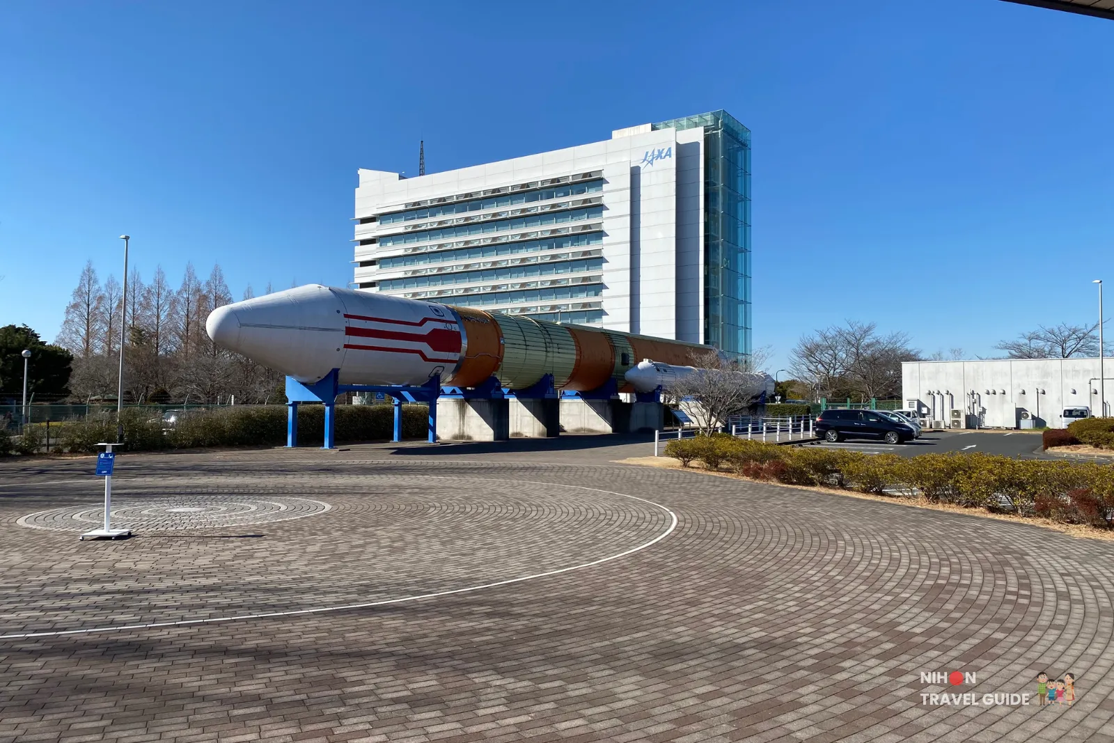 Viewing area with camera stand in the foreground facing a full-scale H-II rocket displayed outside the JAXA Tsukuba Space Center main building on a clear day.