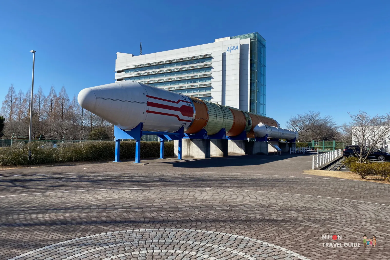 jaxa-tsukuba-space-center-h2-rocket-exterior Full-scale H-II rocket displayed outdoors in front of the JAXA Tsukuba Space Center main building on a clear sunny day.