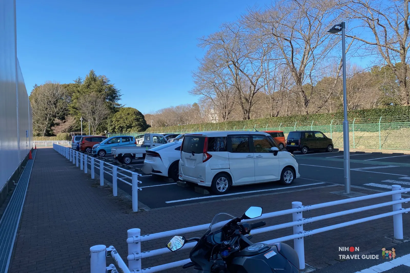 jaxa-tsukuba-space-center-parking-lot Outdoor parking lot at JAXA Tsukuba Space Center with cars and a motorcycle parked beside white railings on a clear blue-sky day.