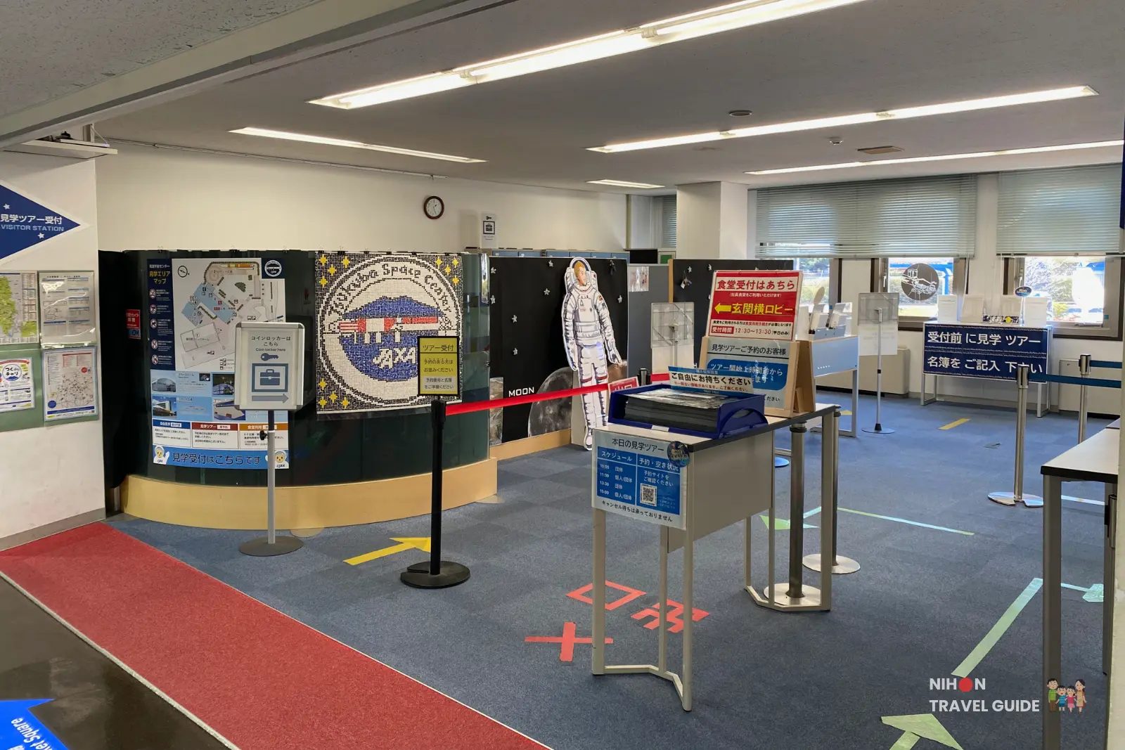 jaxa-tsukuba-space-center-visitor-reception Visitor reception area at JAXA Tsukuba Space Center with tour information signs, a JAXA logo display and an astronaut cutout inside the main building.