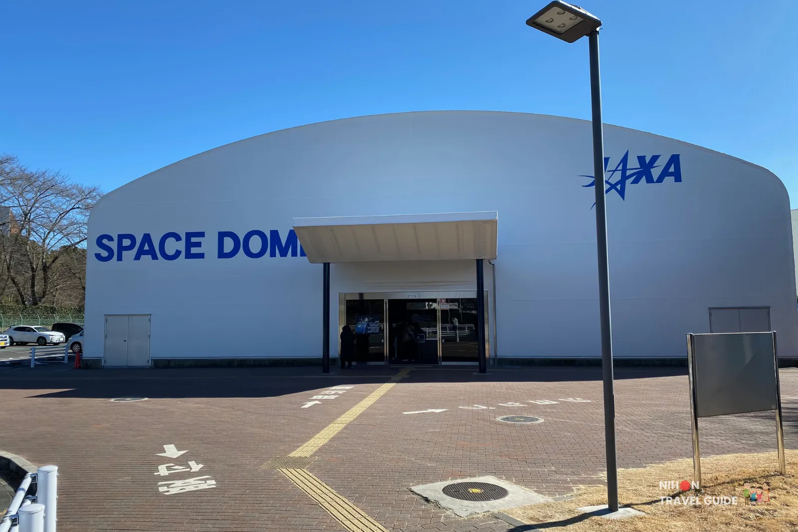 Front view of the JAXA Space Dome exhibition building at Tsukuba Space Center with the main entrance under a small canopy on a clear blue-sky day.