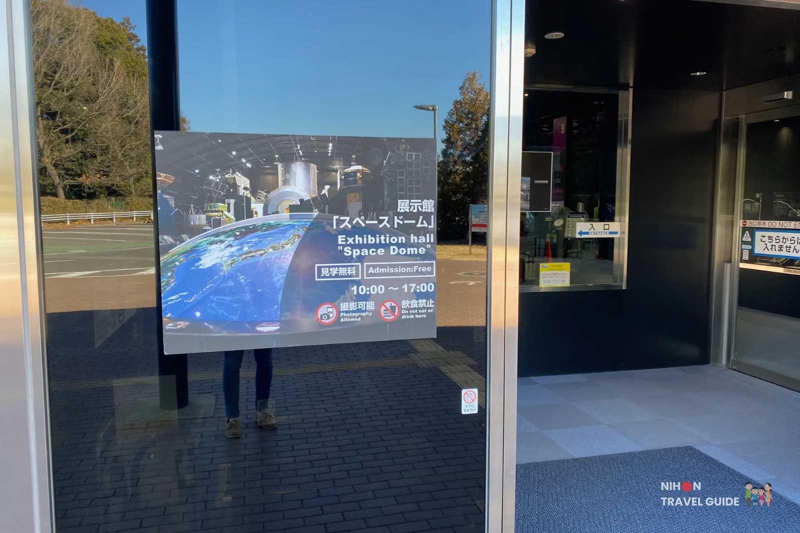 Poster on the entrance glass advertising the Space Dome exhibition hall at JAXA Tsukuba Space Center, noting free admission and opening hours 10:00–17:00.