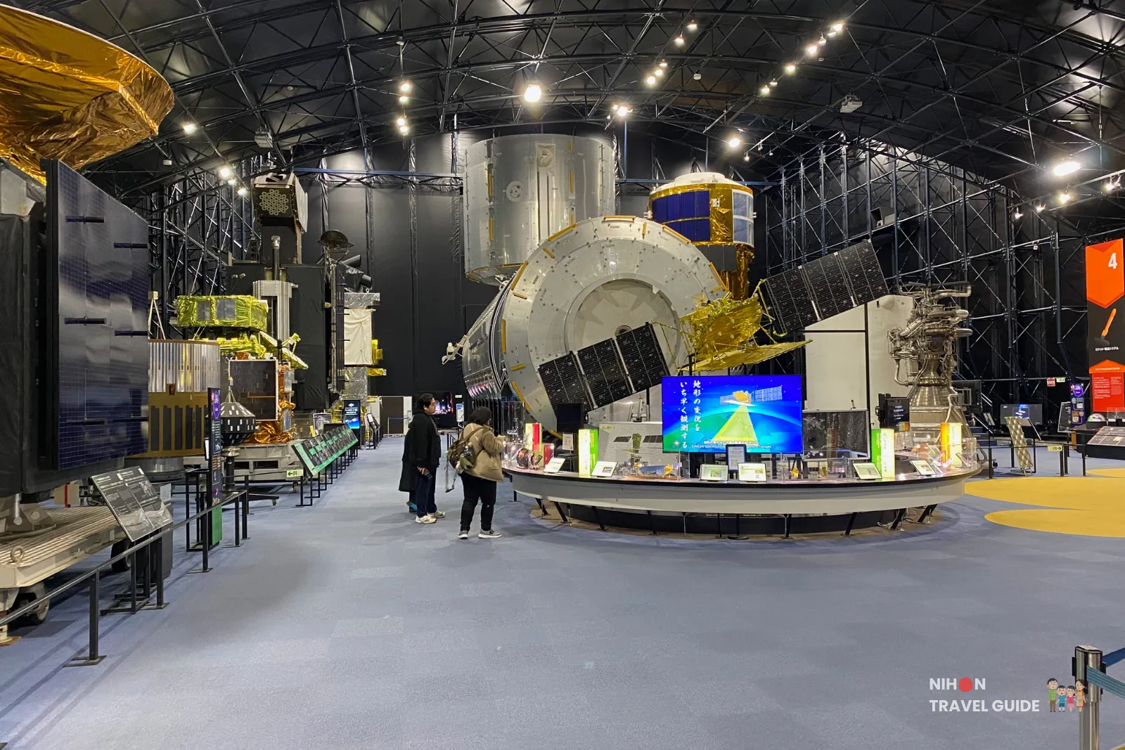 Interior of the Space Dome at JAXA Tsukuba Space Center with large satellite and spacecraft models displayed along a wide exhibition hall.