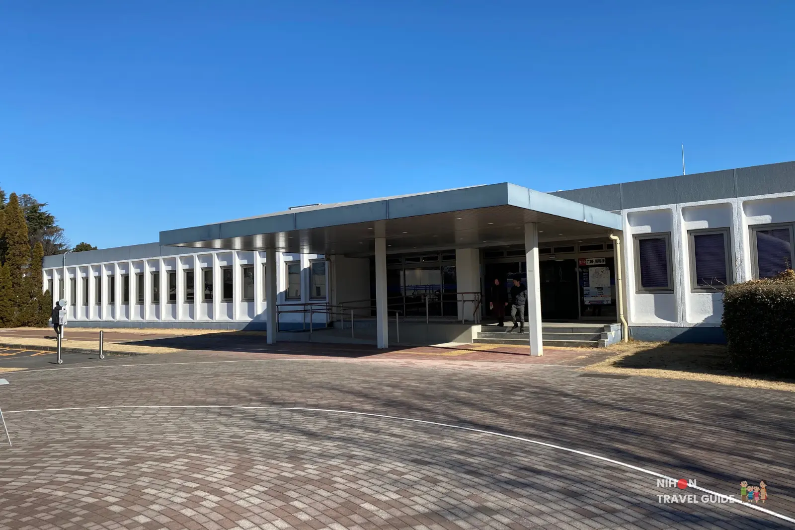 jaxa-tsukuba-visitor-building-exterior-entrance Exterior of the low white visitor building next to the Space Dome at JAXA Tsukuba Space Center, with a covered entrance and paved plaza in front.