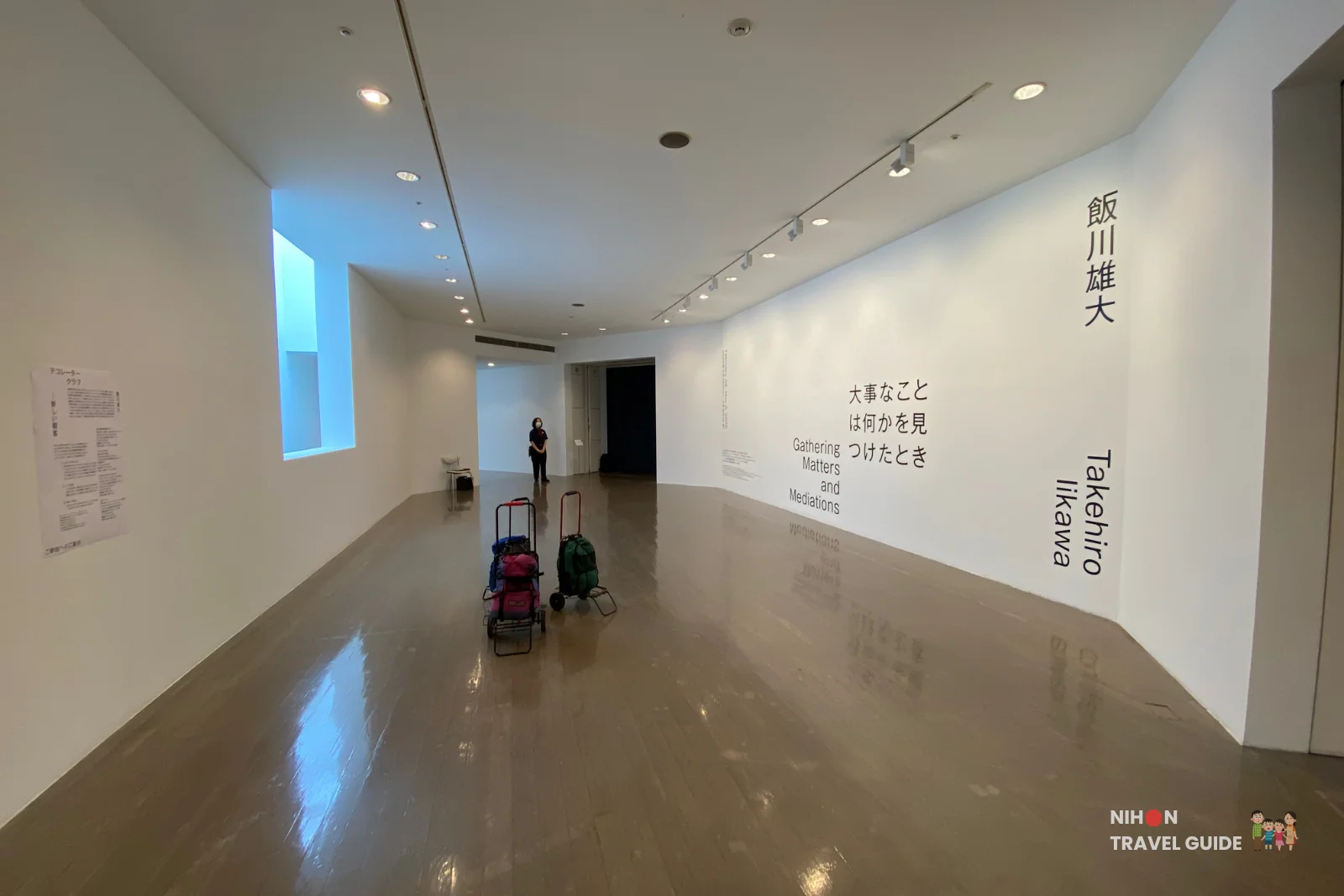 Long view of the bright white gallery space in Takehiro Iikawa’s “Decorator Crab — Make Space, Use Space” exhibition at Art Tower Mito, with wall text and luggage carts on the polished floor.