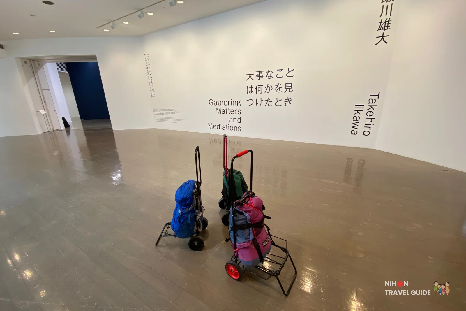 Spacious gallery view of the Takehiro Iikawa "Gathering Matters and Mediations" exhibition at Art Tower Mito, with wall text, luggage carts in the foreground, and polished reflective floor.