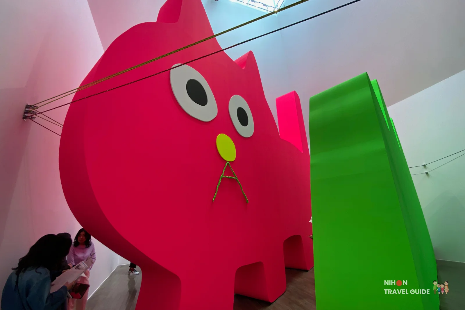Large bright pink cat sculpture with green elements in the Takehiro Iikawa "Gathering Matters and Mediations" exhibition at Art Tower Mito, visitors viewing the artwork.