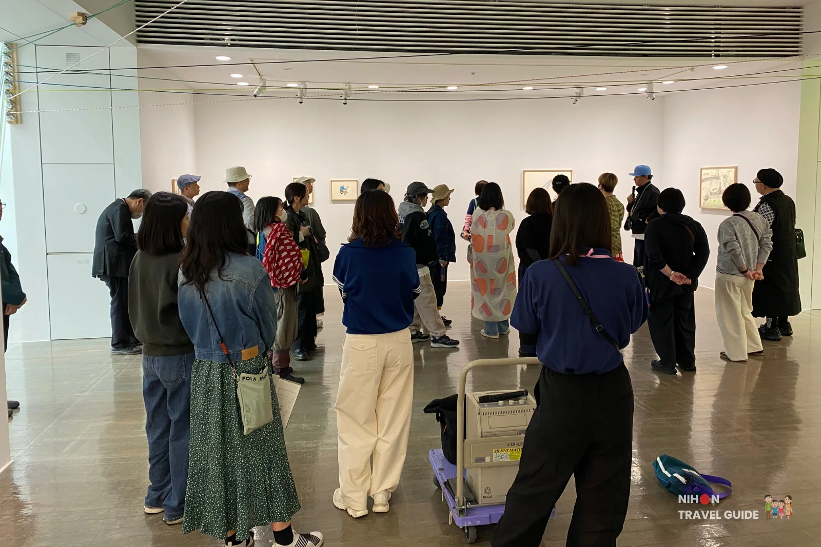 Takehiro Iikawa personally giving a guided tour of his exhibition to a group of visitors at Art Tower Mito, with attendees listening attentively in the gallery space.