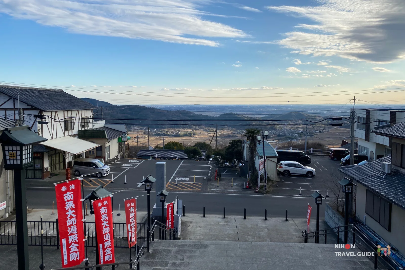 View down the road below Tsukubasan Shrine, with small roadside shops, parking areas, and wide plains visible in the distance from the shrine steps.