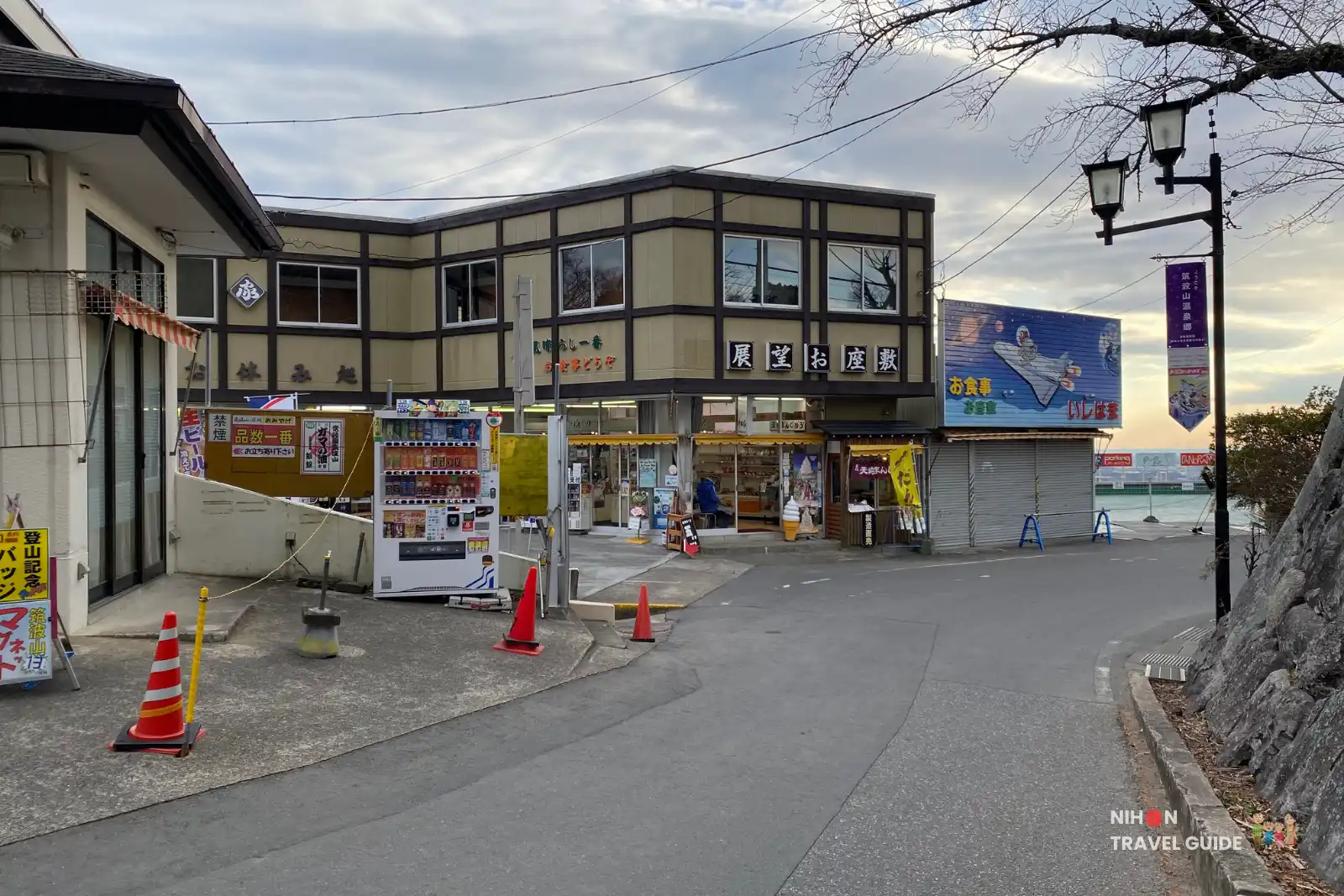 Quiet street near Tsukubasan Shrine lined with souvenir shops, vending machines, and parking areas overlooking the plain below.