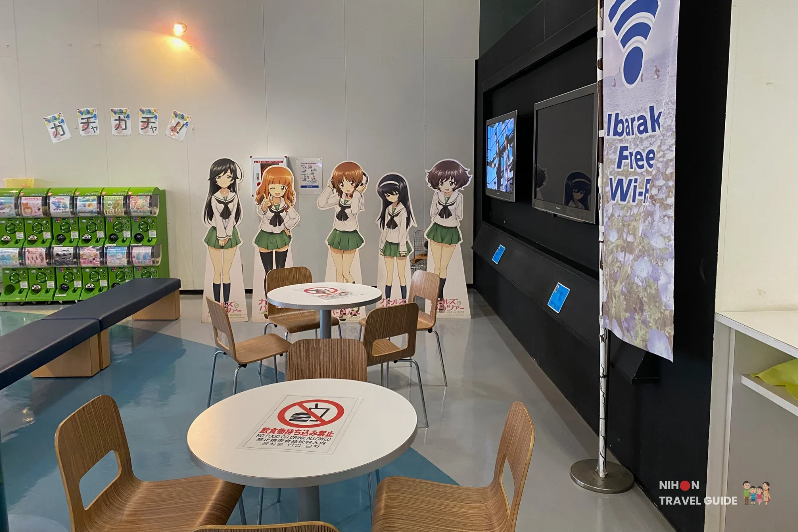 Relaxing seating area at Oarai Marine Tower featuring life-size Girls und Panzer anime character cutouts, round tables, chairs, green gashapon machines, and Ibaraki Free Wi-Fi banner, Japan