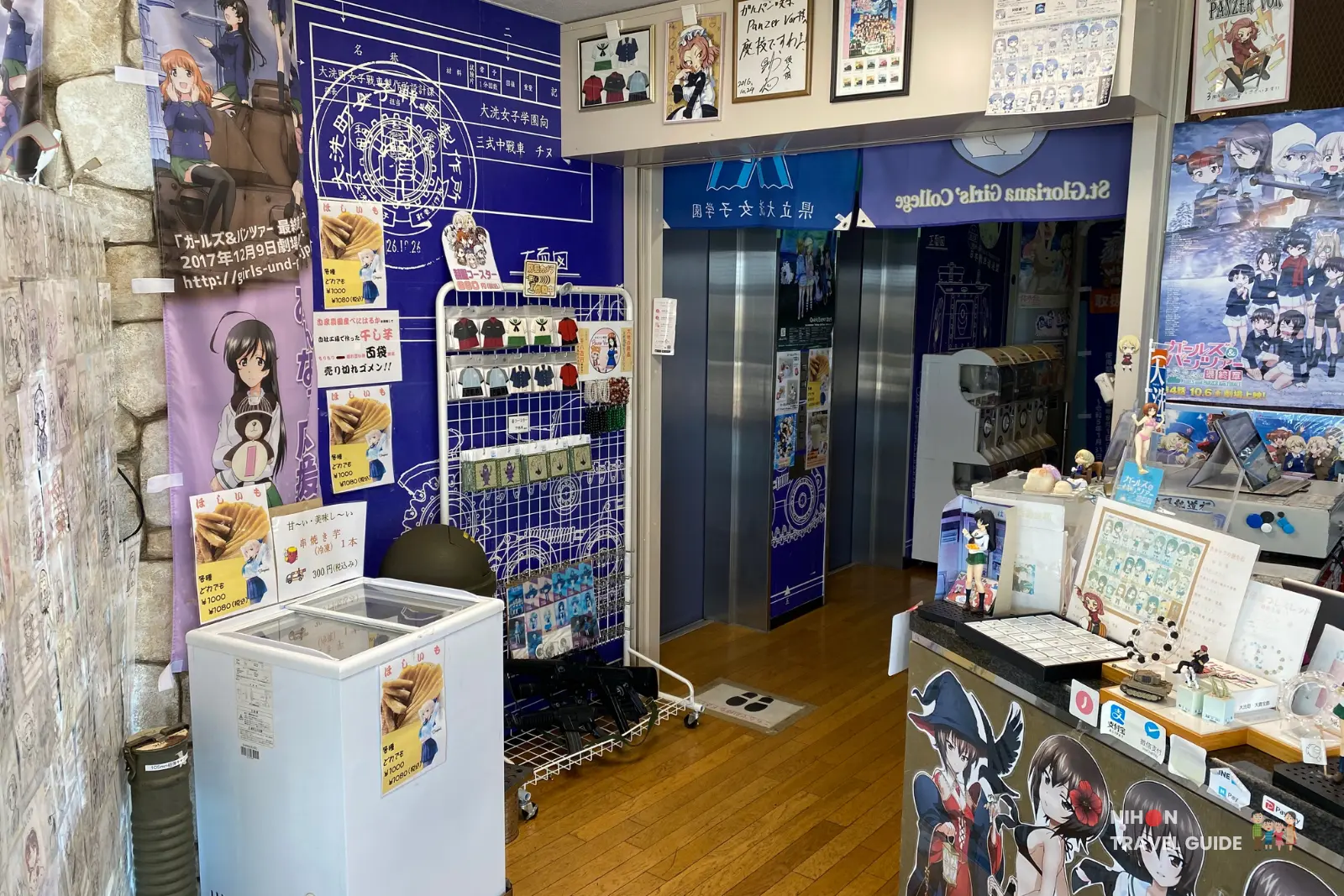 Overview of Café Panzer Vor at Oarai Marine Tower showing the service counter, elevator, blue Girls und Panzer themed walls, merchandise displays, and fan art wall, Ibaraki Prefecture, Japan