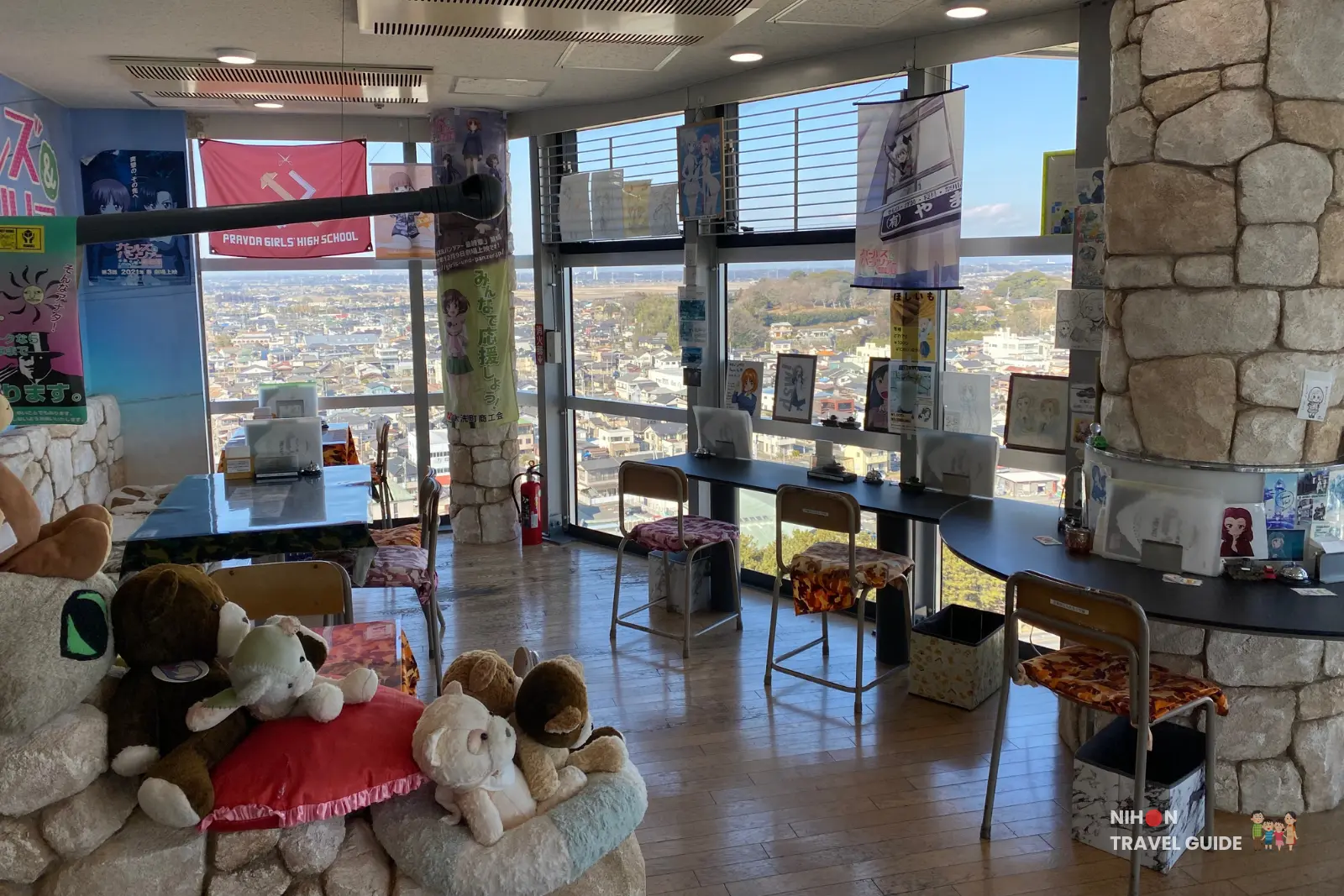 Cozy lounge area of Café Panzer Vor on the second floor of Oarai Marine Tower, featuring Girls und Panzer plush toys, Pravda Girls High School banner, and panoramic windows overlooking the town and sea, Ibaraki, Japan