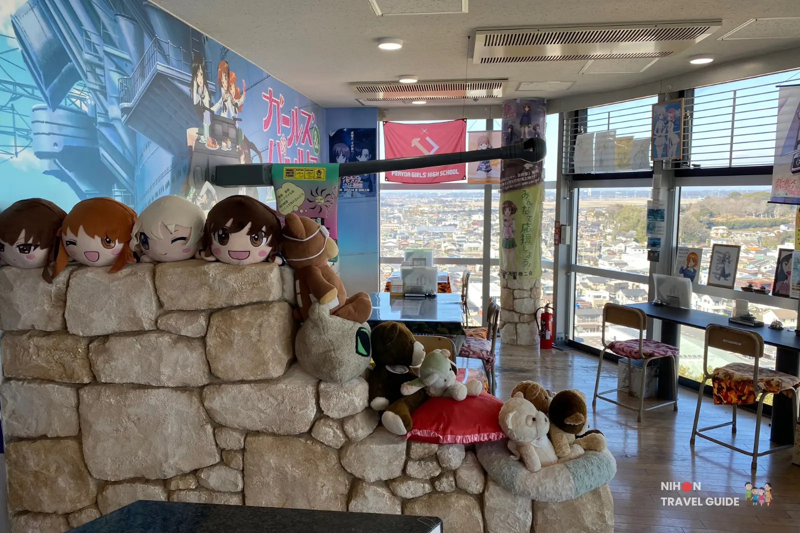 Adorable Girls und Panzer character plush toys lined up on a stone wall inside Café Panzer Vor at Oarai Marine Tower, with large windows overlooking the town and sea in the background, Japan