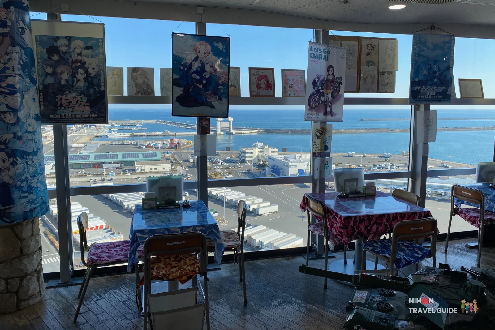 Seating area inside Café Panzer Vor at Oarai Marine Tower offering a stunning panoramic view of the Pacific Ocean and Oarai port, decorated with Girls und Panzer anime posters and hanging artwork, Ibaraki Prefecture, Japan
