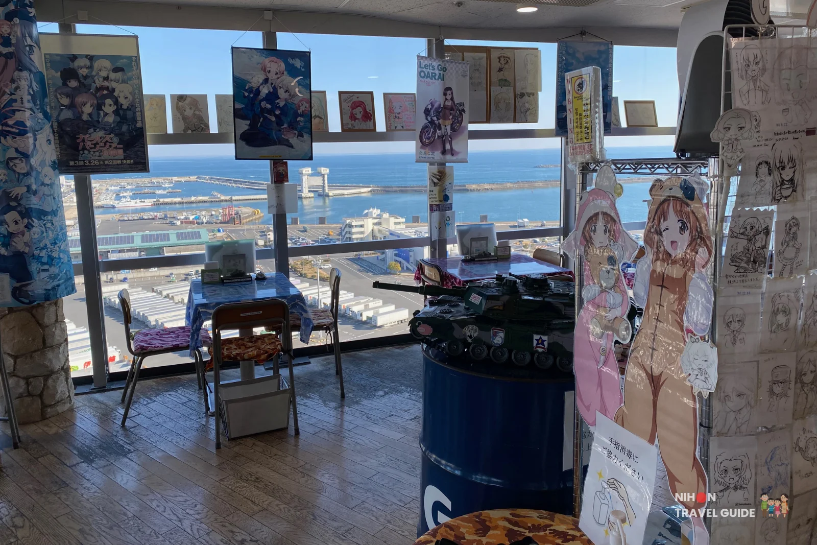 Café Panzer Vor interior at Oarai Marine Tower with Girls und Panzer character cutouts, a model tank on a blue barrel, and breathtaking views of the ocean and harbor through large windows, Ibaraki, Japan