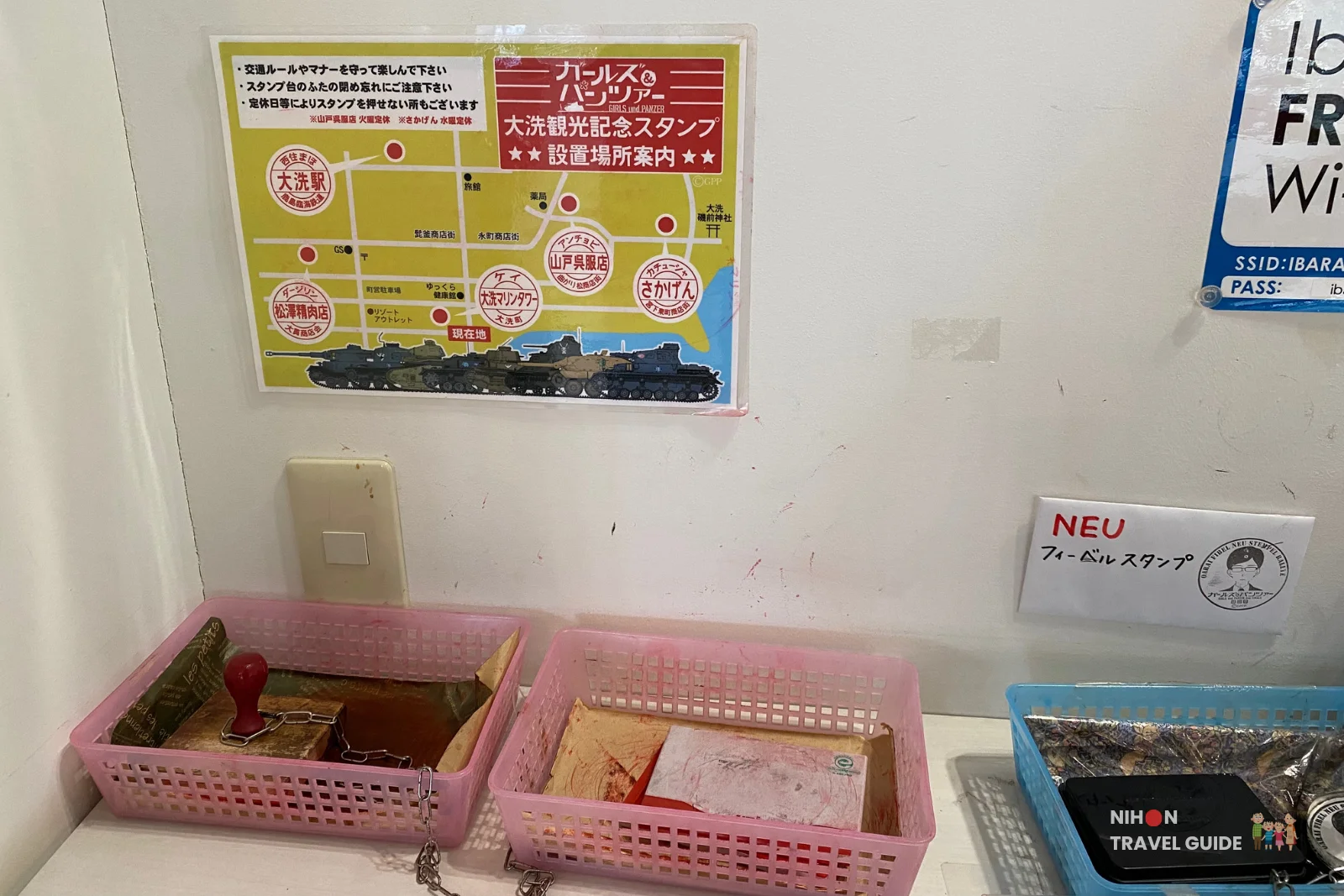 Close-up of the Girls und Panzer stamp rally station at Oarai Marine Tower, showing wooden stamp pads in pink baskets and a colorful map poster highlighting stamp locations including Oarai Marine Tower, Ibaraki, Japan