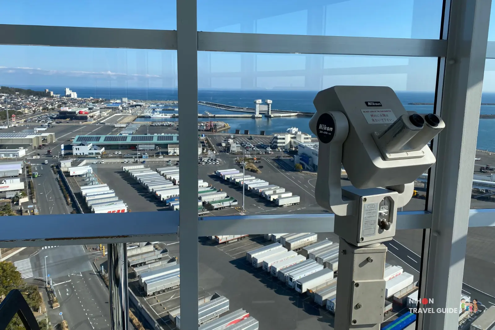 Nikon coin-operated binoculars on the observation deck of Oarai Marine Tower framing the east view of the Pacific Ocean, Oarai port, and container terminal on a bright sunny day, Ibaraki, Japan