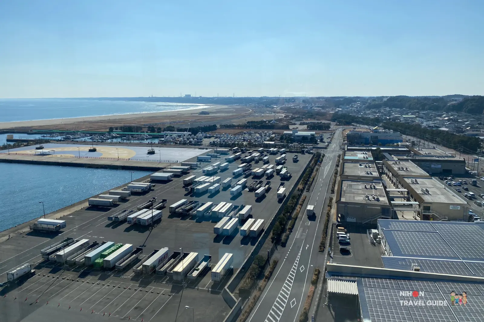 Panoramic south view from Oarai Marine Tower observation deck showing the busy Oarai port, container yard with rows of trucks and trailers, marina, Oarai Sun beach, and distant town under a clear blue sky, Ibaraki Prefecture, Japan