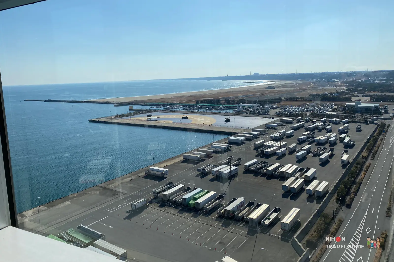 Panoramic south view from Oarai Marine Tower observation deck showing the busy Oarai port, container yard with rows of trucks and trailers, marina, Oarai Sun beach, and distant town under a clear blue sky, Ibaraki Prefecture, Japan
