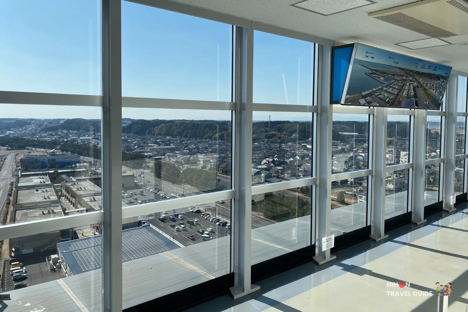 Bright interior of the observation deck at Oarai Marine Tower with large floor-to-ceiling windows offering sweeping south views over the town, port, and forested hills, plus an informational display panel, Ibaraki, Japan