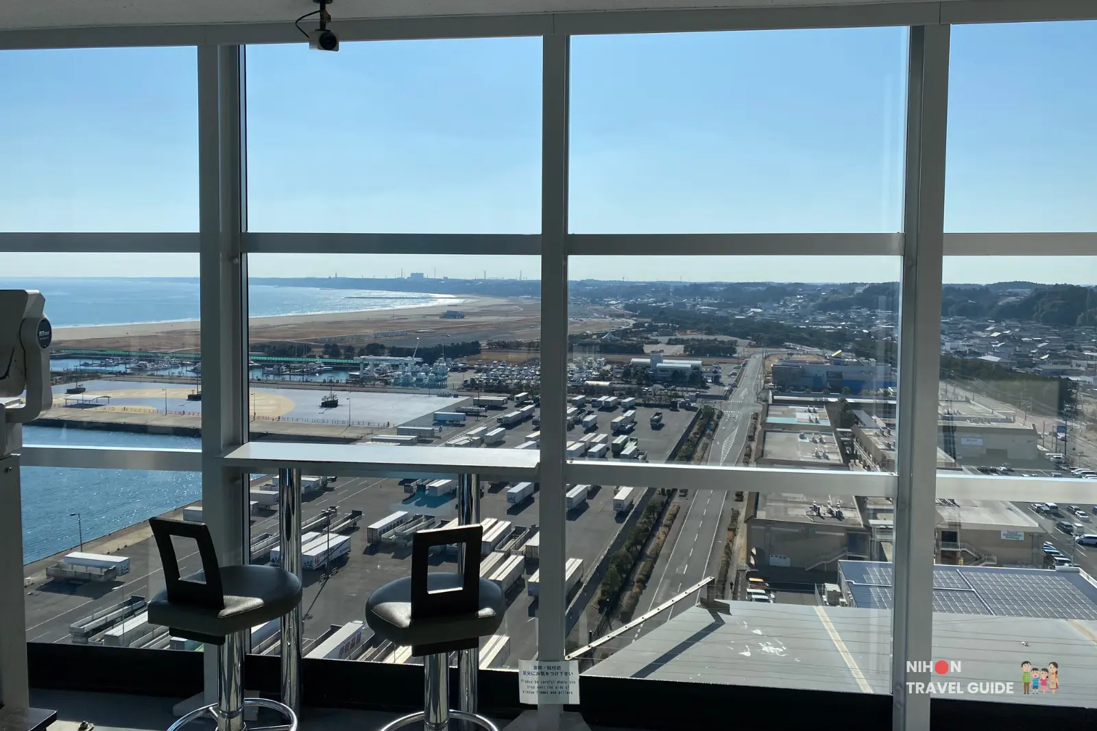 Comfortable seating area with high stools at the south-facing windows of Oarai Marine Tower observation deck, overlooking the Pacific Ocean, harbor, container yard, and sandy beach on a clear day, Ibaraki Prefecture, Japan.