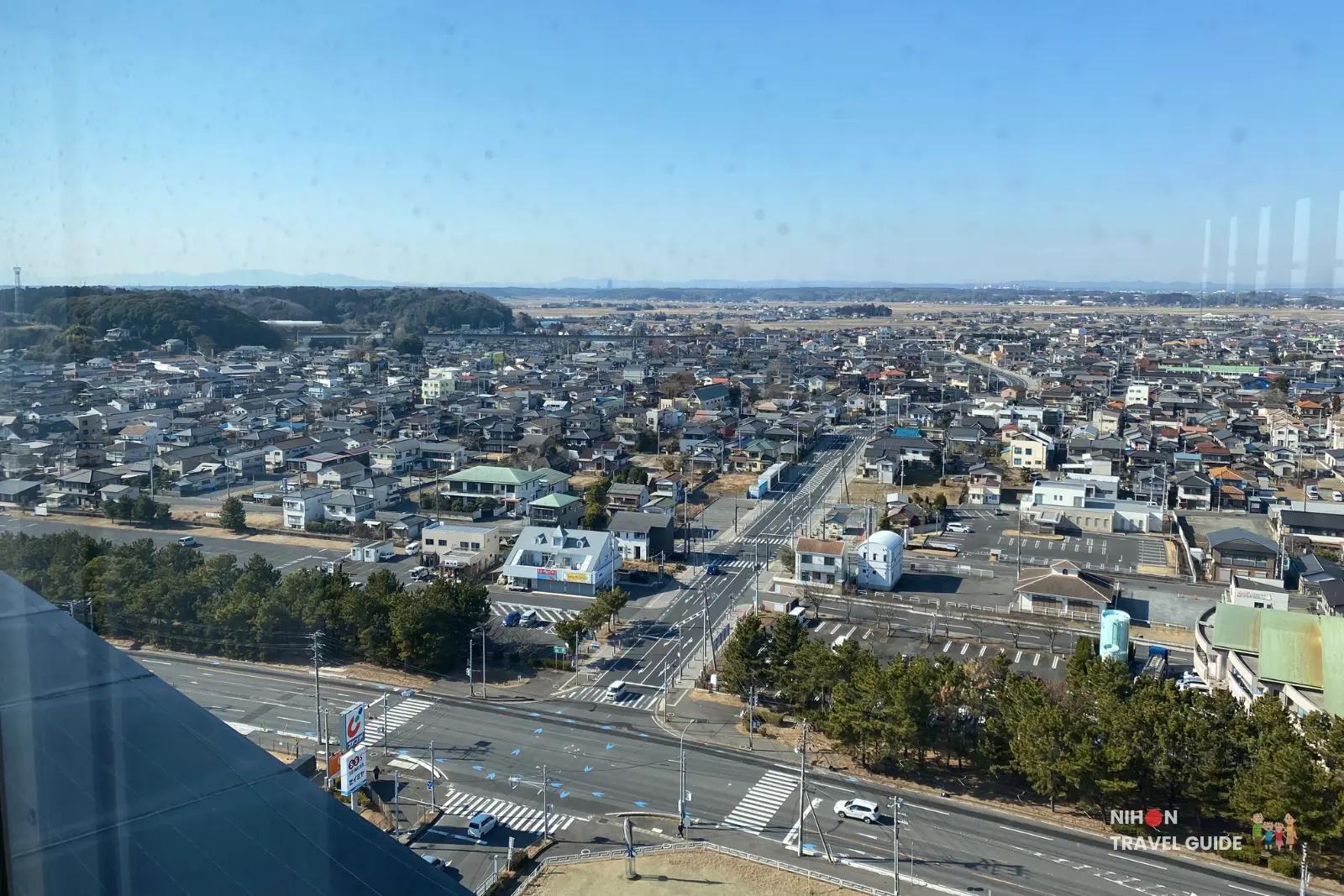 Southwest view from the observation deck of Oarai Marine Tower showing a wide expanse of the town, a prominent white building, major highway with traffic, pine groves, and mountains visible in the far distance, Ibaraki Prefecture, Japan