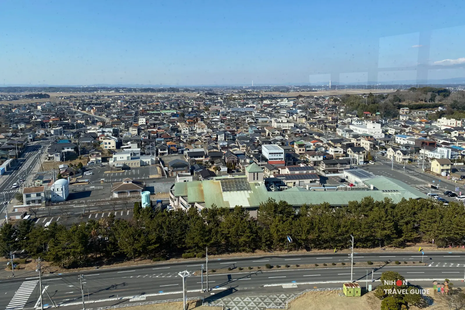 Southwest panoramic view from Oarai Marine Tower observation deck showing the dense residential townscape of Oarai, major roads, a large green-roofed building, pine trees, and distant mountains on a clear sunny day, Ibaraki Prefecture, Japan