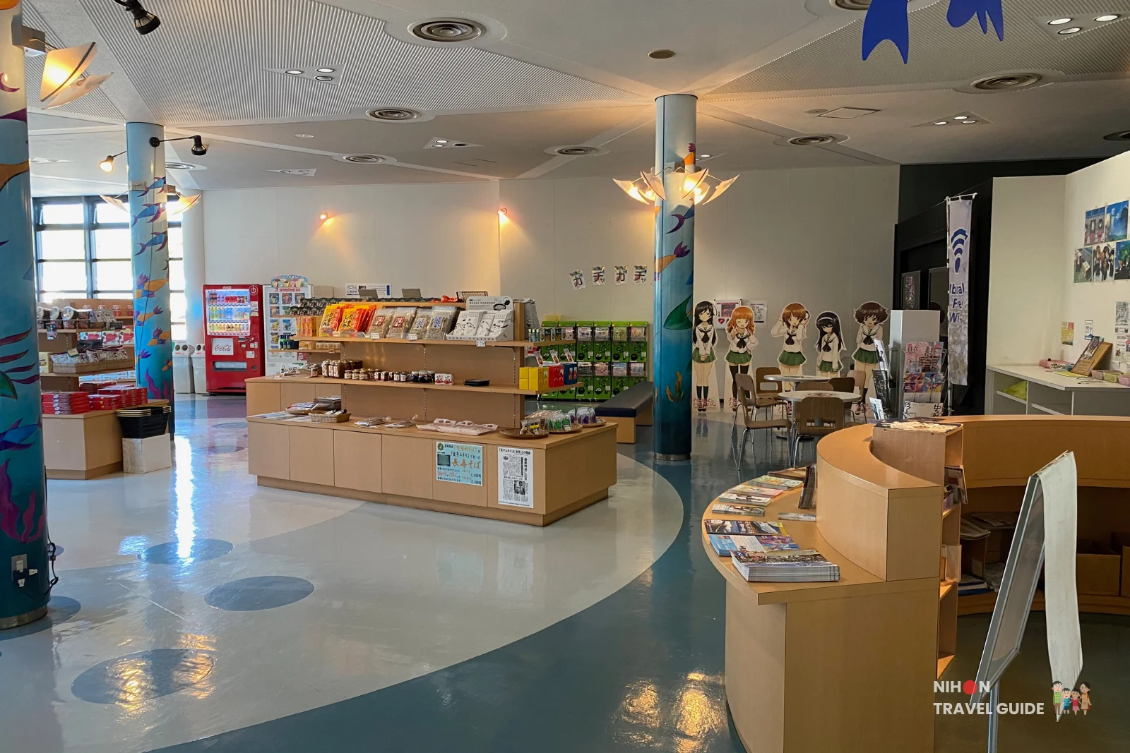 Wide view of the souvenir shop at Oarai Marine Tower showing wooden display counters, a curved information desk, anime character cutouts, a red vending machine, and decorative sea-themed pillars, Oarai, Ibaraki, Japan