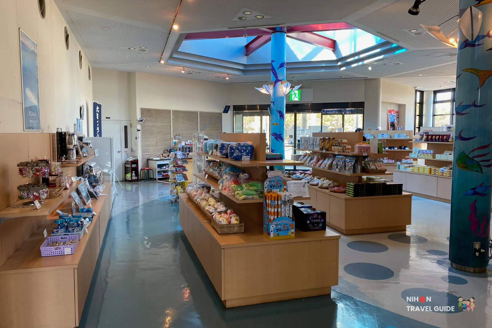 Bright interior of the souvenir shop at Oarai Marine Tower, featuring wooden display shelves stocked with plush toys, snacks, and marine-themed goods, colorful dolphin-painted pillars, and a large skylight, Ibaraki Prefecture, Japan