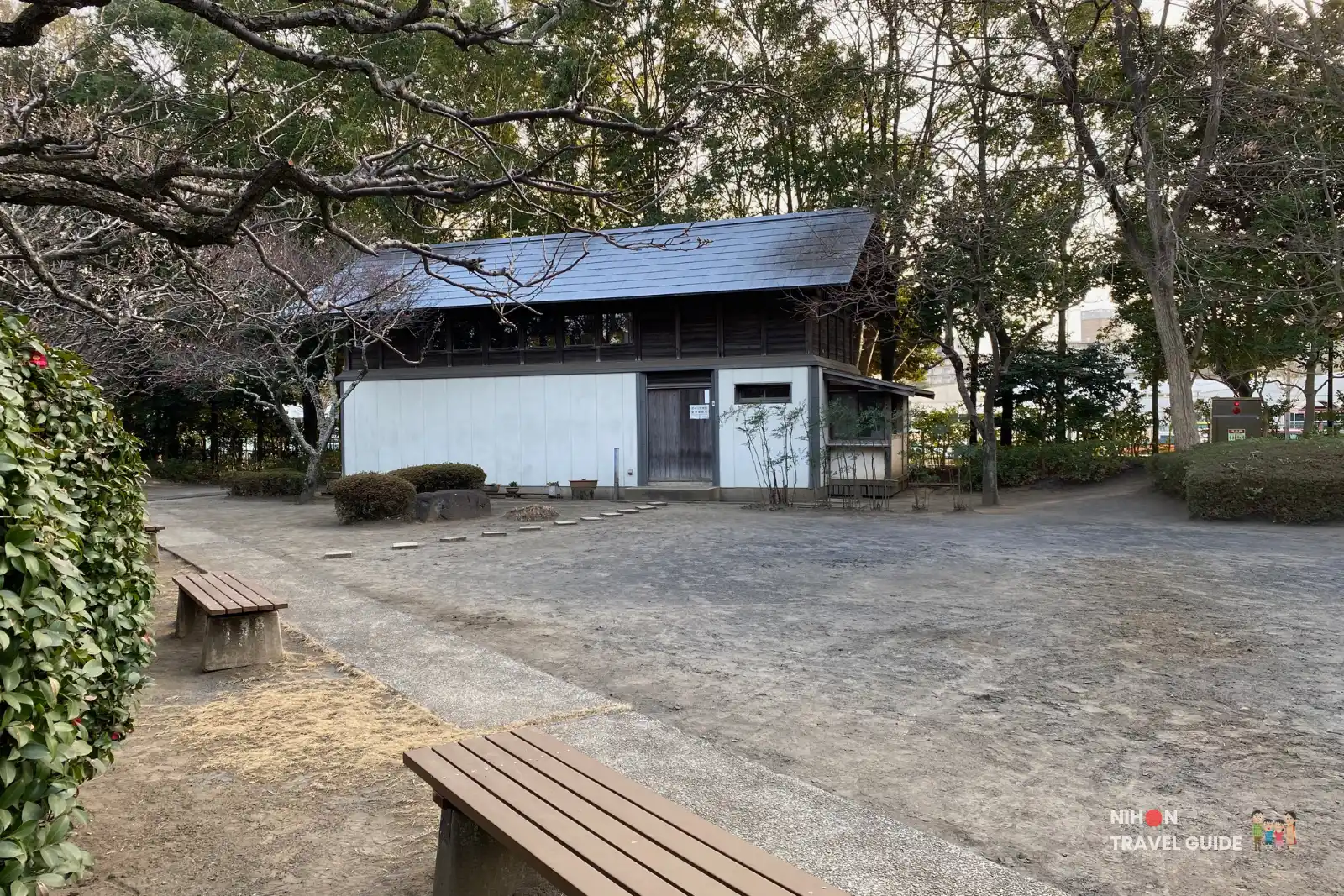 Smaller traditional wooden building with a blue-tiled roof at Sakura Minka-en. The open courtyard and surrounding trees create a peaceful historic atmosphere.